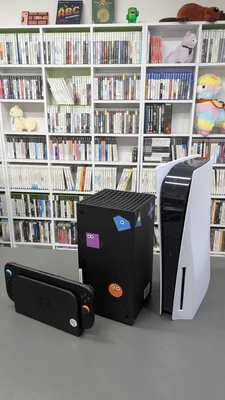 Trois consoles de jeux de generation actuelle sont placees sur une table grise devant une grande etagere blanche remplie de jeux video. A gauche se trouve une Nintendo Switch 2 avec ses manettes attachees, au centre une Xbox Series X noire avec des autocollants colores, et a droite une PlayStation 5 blanche placee verticalement. En arriere-plan, de nombreuses boites de jeux sont rangees avec soin ainsi que quelques figurines decoratives et peluches.