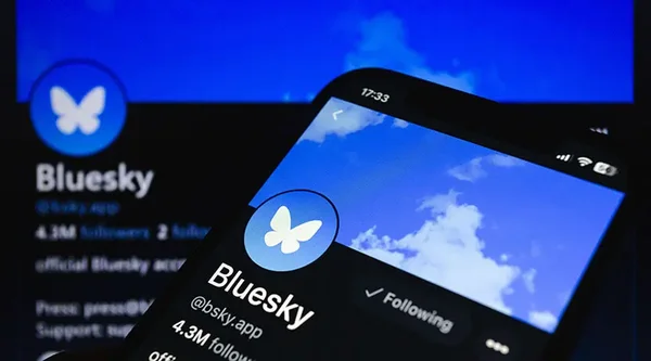 Smartphone affichant l'application Bluesky avec un écran d'ordinateur en arrière-plan.
