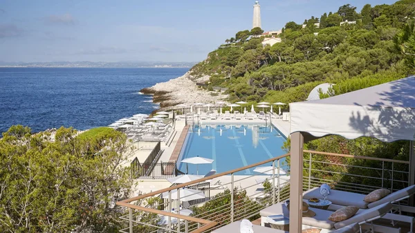 Complexe hôtelier de luxe avec piscine surplombant la mer, phare et terrasse ombragée.