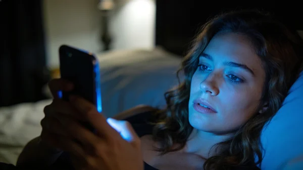 Femme regardant son téléphone dans son lit le soir, écran éclairant son visage, chambre tamisée