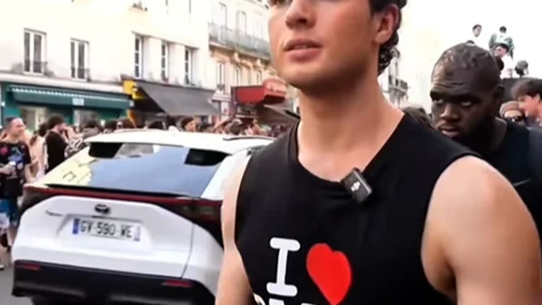 Clavicular en débardeur noir dans une rue animée, avec une voiture blanche et des immeubles en arrière-plan.
