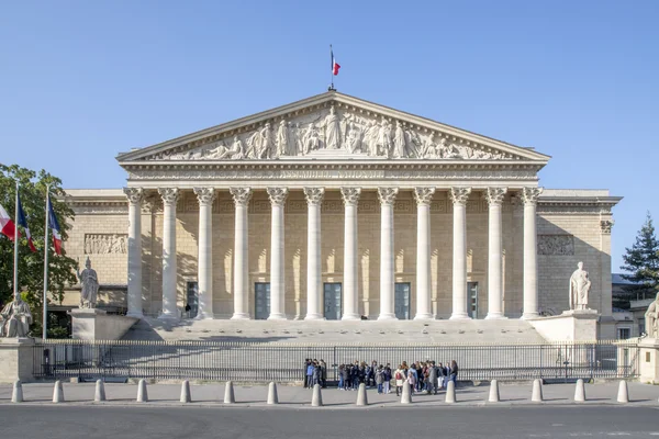 Le Palais Bourbon sous un ciel bleu, avec des personnes rassemblées devant l'entrée.