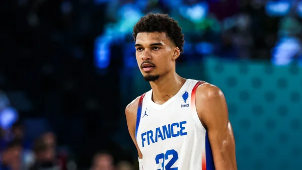 Un plan moyen d'un joueur de basket-ball masculin portant un maillot blanc avec 'FRANCE' et le numéro 32 imprimés sur le devant, debout contre un fond flou de lumières bleues d'arène.