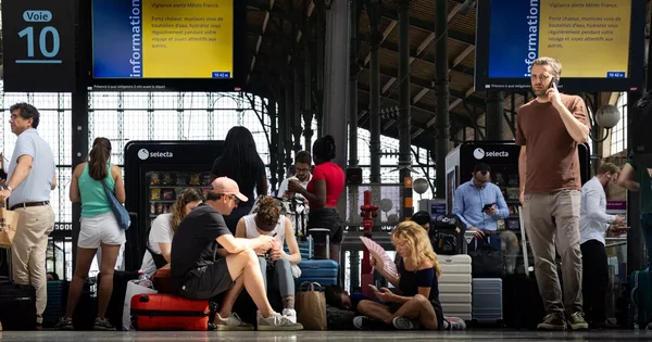 Voyageurs en attente sur un quai de gare, avec bagages et écrans d'information affichant des alertes.