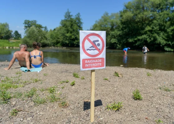Panneau 'Baignade Interdite' sur une rivière, avec des baigneurs en arrière-plan.