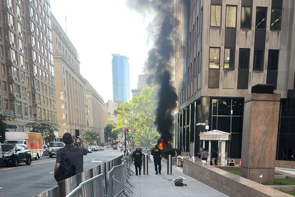 Un incendie sur le trottoir devant un immeuble fédéral à New York, avec des policiers se dirigeant vers les lieux et un témoin filmant la scène.