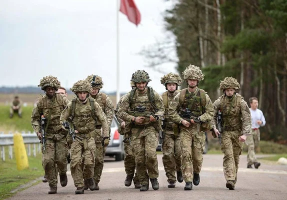 Soldats britanniques en marche lors d'un exercice de mobilisation, avec un drapeau rouge en arrière-plan.