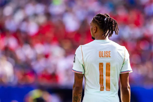 Un plan de dos d'un joueur de football masculin avec des dreadlocks portant un maillot blanc avec le nom 'OLISE' et le numéro '11' en orange, debout sur un terrain avec une foule floue du stade en arrière-plan.