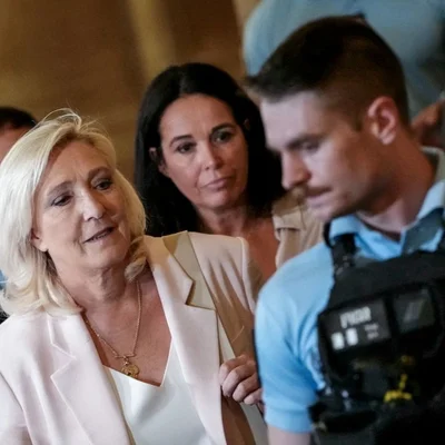 Marine Le Pen, veste blanche et collier doré, devant un agent de sécurité.