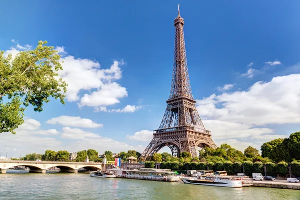 La tour Eiffel vue depuis la Seine, avec des bateaux-mouches amarrés et un pont de pierre à gauche.