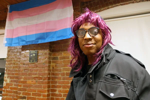 Personne aux cheveux violets devant un mur de briques, un drapeau transgenre en arrière-plan.