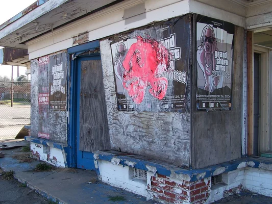 Photographie d'une station-service abandonnée aux murs tagués, avec une affiche promotionnelle du jeu vidéo Grand Theft Auto: San Andreas visible sur la façade délabrée.
