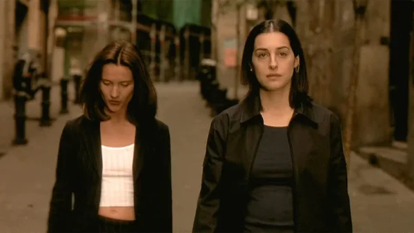 Deux femmes avançant dans une ruelle sombre, l'une vêtue de blanc et de noir, l'autre en veste noire.