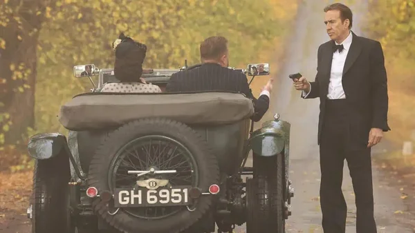 Nicolas Cage en costume noir sur une route, pointant un pistolet vers une voiture décapotable verte dans une scène du tournage de Fortitude, thriller d'espionnage de la Seconde Guerre mondiale réalisé par Simon West.