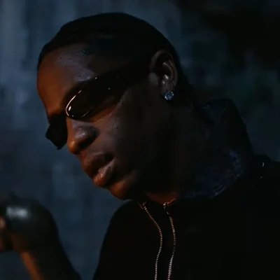 Portrait en gros plan de Travis Scott portant des lunettes de soleil et une veste noire à fermeture éclair, dans une ambiance sombre, extrait du clip 'When I'm Home'.