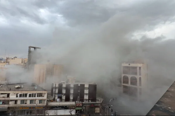 Un épais nuage de fumée et de poussière s'élève au-dessus d'une rue de Téhéran après une frappe, des bâtiments à plusieurs étages partiellement visibles sous un ciel couvert.