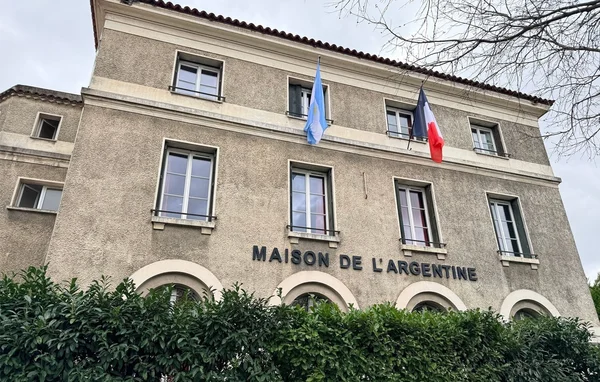 Vue en contre-plongée de la Maison de l'Argentine à la Cité internationale universitaire de Paris, bâtiment en stuc beige avec les drapeaux argentin et français hissés sur la façade.