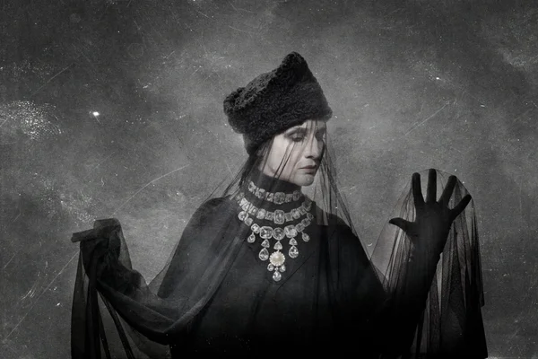 Portrait en noir et blanc de Claire Diterzi portant un chapeau de fourrure sombre, un voile transparent et un collier de diamants élaboré, devant un fond texturé avec des rayures visibles.