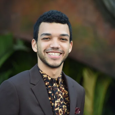 Portrait de l'acteur Justice Smith, souriant avec une barbe légère, portant un blazer sombre sur une chemise à motif, pour la saison 2 de Heated Rivalry