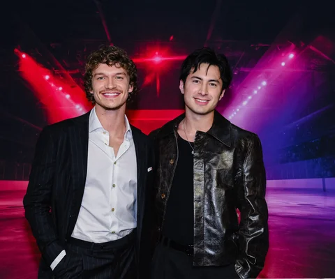 Les deux acteurs principaux de la saison 2 de Heated Rivalry posent côte à côte devant un fond sombre éclairé de lumières rouges et violettes, l'un en costume à rayures, l'autre en blouson de cuir noir.
