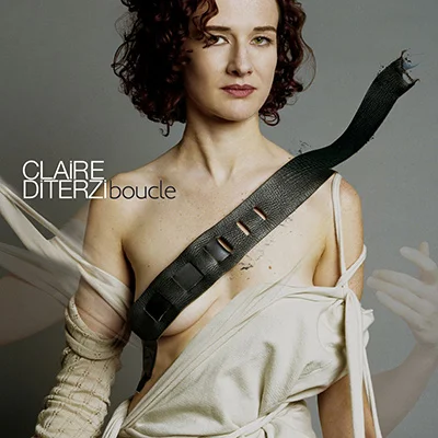 Portrait de Claire Diterzi, cheveux bouclés courts, vêtue d'une draperie blanche avec une lanière de cuir noir sur la poitrine, pour son album Boucle (2006)