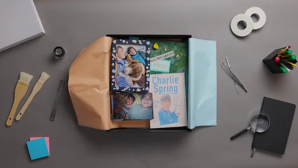 Vue de dessus d'une boîte noire ouverte contenant des objets souvenirs de la série Heartstopper, avec un livre intitulé 'Charlie Spring', des photos de deux jeunes hommes et un chien, et un carnet vert marqué 'Nick and Charlie's Memories', entourés de fournitures de bureau sur fond gris.