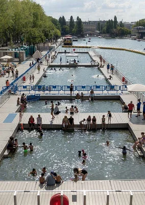Baignade en Seine à Paris : vue aérienne d'un complexe de piscines flottantes sur la rivière, avec des bassins rectangulaires occupés par des nageurs sur des pontons en bois, arbres et immeubles en fond.