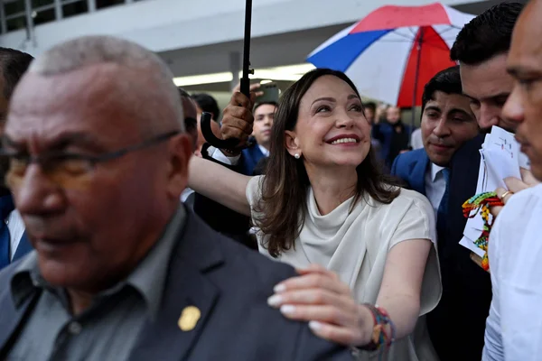 María Corina Machado, cheveux bruns aux épaules, portant un haut blanc et des bracelets colorés, souriant vers le haut dans une foule en tenant un manche de parapluie, avec un homme aux lunettes au premier plan.