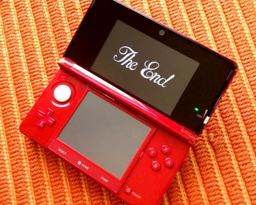 Un Nintendo 3DS XL rouge ouvert sur une surface orange, affichant le texte "The End" en police blanche et cursive sur son écran supérieur, montrant la fin du jeu Dragon Quest VII pour la Nintendo 3DS.