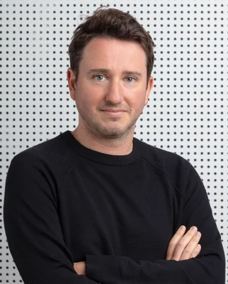 Portrait en studio de l'économiste Gabriel Zucman, cheveux bruns courts et légère barbe, vêtu d'un pull noir à col rond les bras croisés devant un panneau blanc perforé