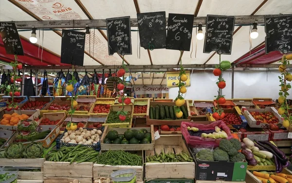 Étal d'un marché alimentaire découvert à Paris présentant des cageots en bois de légumes et fruits frais, artichauts, poivrons, concombres, champignons, brocolis, tomates et oranges, sous des ardoises de prix manuscrites