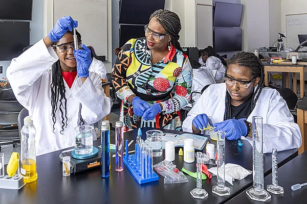 Trois jeunes femmes en blouse blanche, lunettes de protection et gants bleus réalisant des expériences de chimie en laboratoire avec béchers, éprouvettes et agitateur magnétique