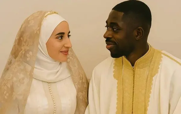 Ousmane Dembélé et Rima Edbouche en élégantes tenues traditionnelles blanches, la femme en hijab blanc avec voile en dentelle, se regardant tendrement sur fond neutre