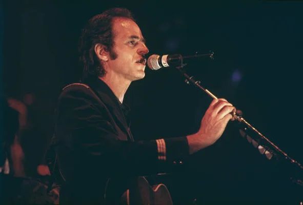 Jean-Jacques Goldman en concert à Besançon, portant un costume sombre et chantant au micro tout en jouant de la guitare acoustique