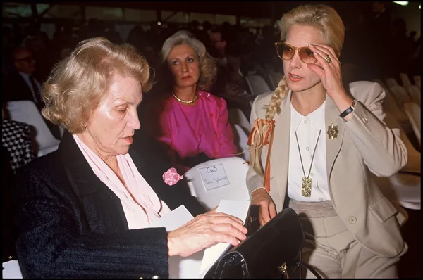 Sylvie Vartan et sa mère assises dans l'audience d'un défilé Chanel, l'une en blouson sombre à foulard rose, l'autre en tailleur beige et lunettes de soleil.