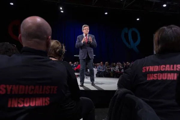 Jean-Luc Mélenchon en costume sur scène prononçant un discours lors de la Convention Nationale des Insoumis, avec des militants en t-shirts noirs « Syndicaliste Insoumis » au premier plan