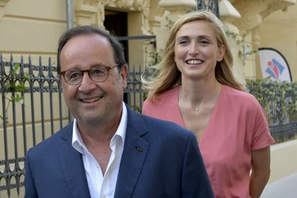François Hollande en costume bleu et lunettes souriant devant un immeuble, avec Julie Gayet en haut rose derrière lui lors de leur rencontre en 2014