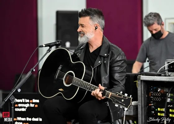 Roch Voisine à barbe grise et veste de cuir noir, souriant en jouant de la guitare acoustique devant un micro sur scène lors de sa tournée Americana Light