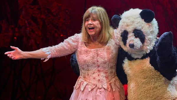 Chantal Goya, en robe rose, debout sur scène aux côtés d'un personnage en costume de panda, devant un décor rouge, lors de son spectacle à Agen Agora.