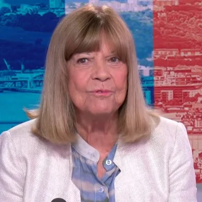 Chantal Goya, cheveux blonds mi-longs avec frange, veste blanche texturée sur une chemise à carreaux bleue, s'exprime face caméra sur un fond coloré lors d'un entretien sur LCI avant sa tournée des 50 ans de carrière.