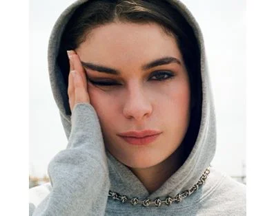 Adèle Castillon en hoodie gris et collier à chaîne, main posée sur le front et clin d’œil, sur la pochette de l’album Plaisir Risque Dépendance.