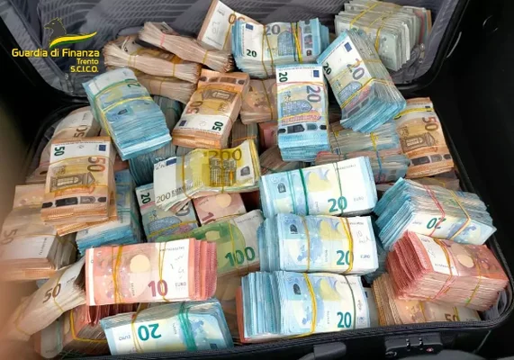 Une valise remplie de liasses de billets en euros de différentes coupures, dont des 10, 20, 50, 100 et 200 euros, avec le logo de la Guardia di Finanza en surimpression.