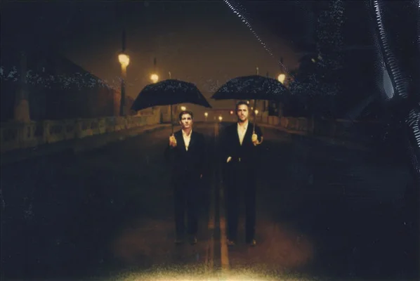 Deux hommes en costume sombre, tenant chacun un parapluie noir, se tiennent dans une rue mouillée et faiblement éclairée la nuit.