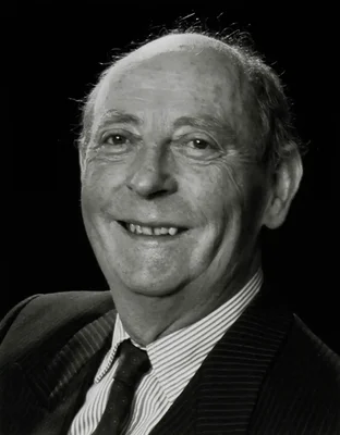 Portrait en noir et blanc de l'écrivain irlandais John McGahern, souriant, vêtu d'un costume rayé, d'une chemise à rayures et d'une cravate sombre sur fond noir.